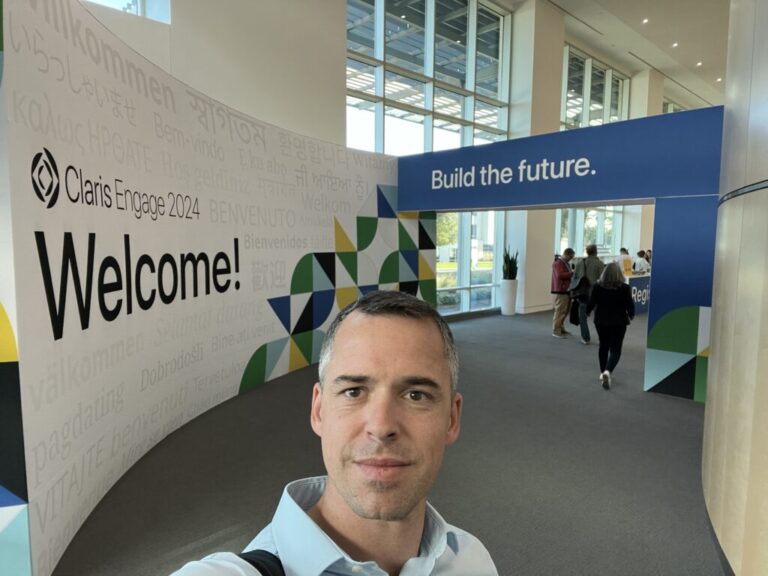 Un homme prend un selfie dans un couloir, devant une enseigne « Claris Engage - Welcome! Build the future. »