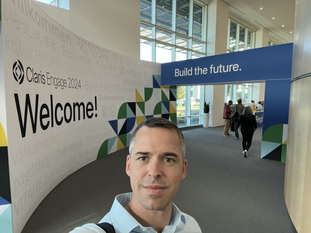 Un homme prend un selfie dans un couloir, devant une enseigne « Claris Engage - Welcome! Build the future. »