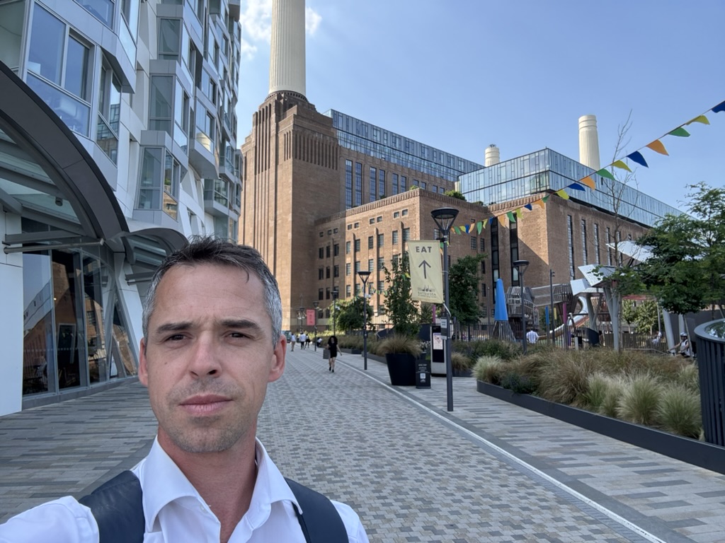Selfie d'un homme dans une rue piétonne, devant un grand ancien bâtiment industriel en briques