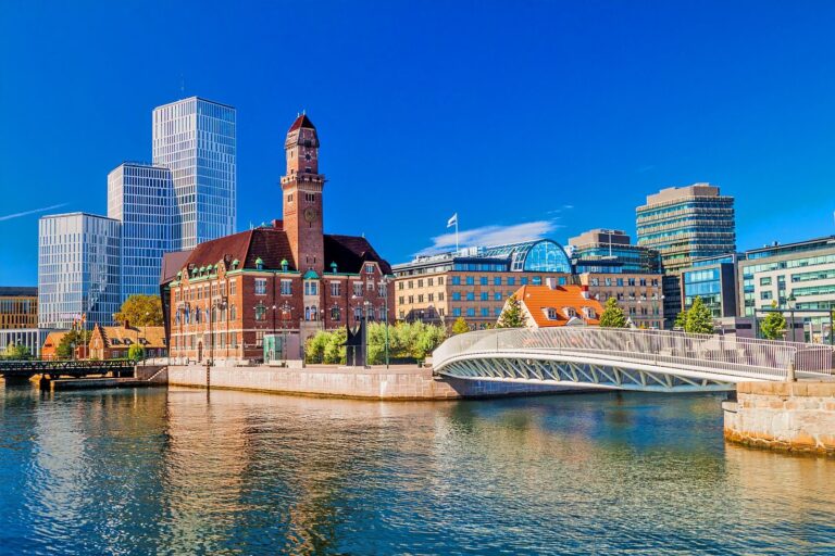 Quai d'un canal avec un bâtiment en briques à tour d'horloge, un pont piéton et des immeubles modernes sous un ciel bleu